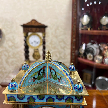 Antique enamel brass quartz clock with intricate design in perfect condition displayed on a wooden table with other clocks in the background